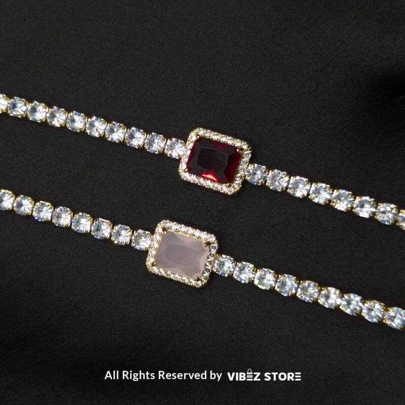 Two crystal-studded gold bracelets with square gemstone centerpieces in ruby red and soft pink on a black fabric background. 