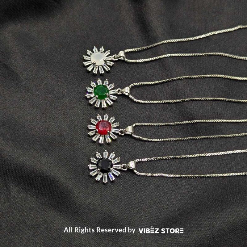 A collection of silver sunburst lockets featuring vibrant gemstones in white, green, red, and black, laid on a black fabric background.

